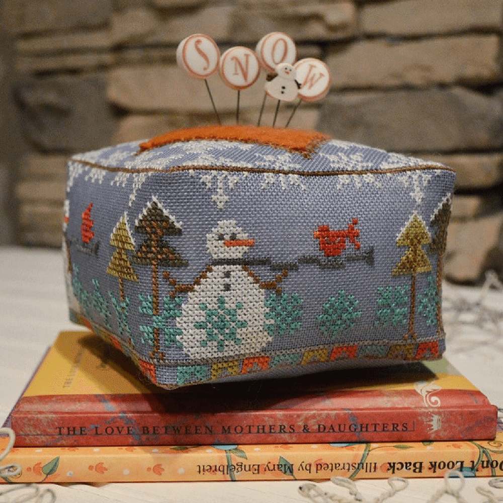 A cross stitch embroidery pincushion kit depicting a snowman and snowy trees on a blue linen background, with a set of pins in the form of snowflakes on top.
Photo by HandsOnDesign.biz [image_link]