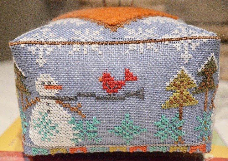 A square cross-stitch cushion with a winter landscape design on a light blue-gray background. The pattern features a white snowman, red birds, evergreen trees, and white snowflakes, with an orange fabric backing visible at the top.
Photo by HandsOnDesign.biz [additional_image_link]