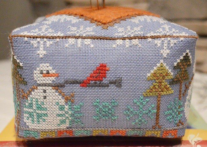 A square cross-stitch needle case featuring a winter scene with a snowman, red bird, evergreen trees, and snowflakes on a light blue-gray background.
Photo by HandsOnDesign.biz [additional_image_link]