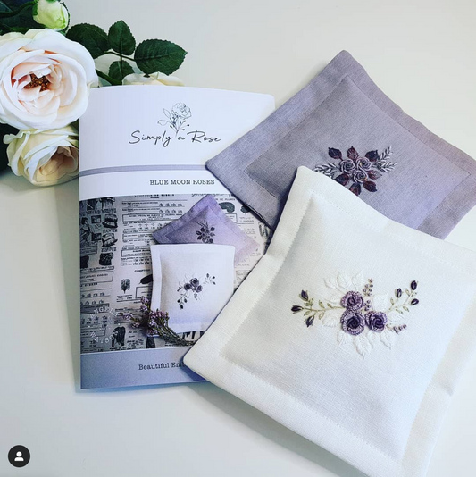 Three embroidered linen doilies in white and soft lavender, arranged on a light surface with a printed booklet and white roses as styling props. The doilies feature floral embroidery in purple, green, and cream.
Photo by Angela Watson [image_link]