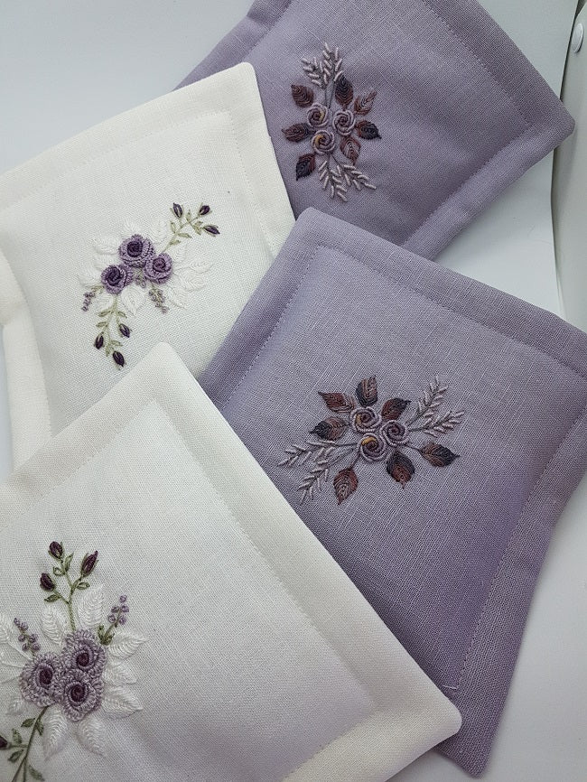 Three square embroidered fabric napkins or pocket squares in white and light lavender, arranged on a white surface. Each piece features floral embroidery in purple, brown, and silver thread.
Photo by Angela Watson [additional_image_link]