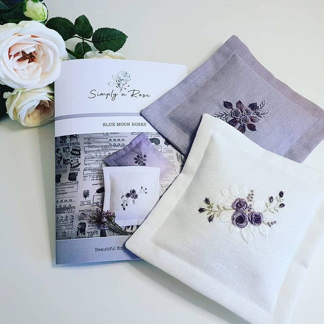 Three embroidered square fabric napkins in white and pale lavender are arranged on a light surface with a printed pattern booklet and white roses. The napkins feature floral embroidery in purple, green, and white.
Photo by Angela Watson [additional_image_link]