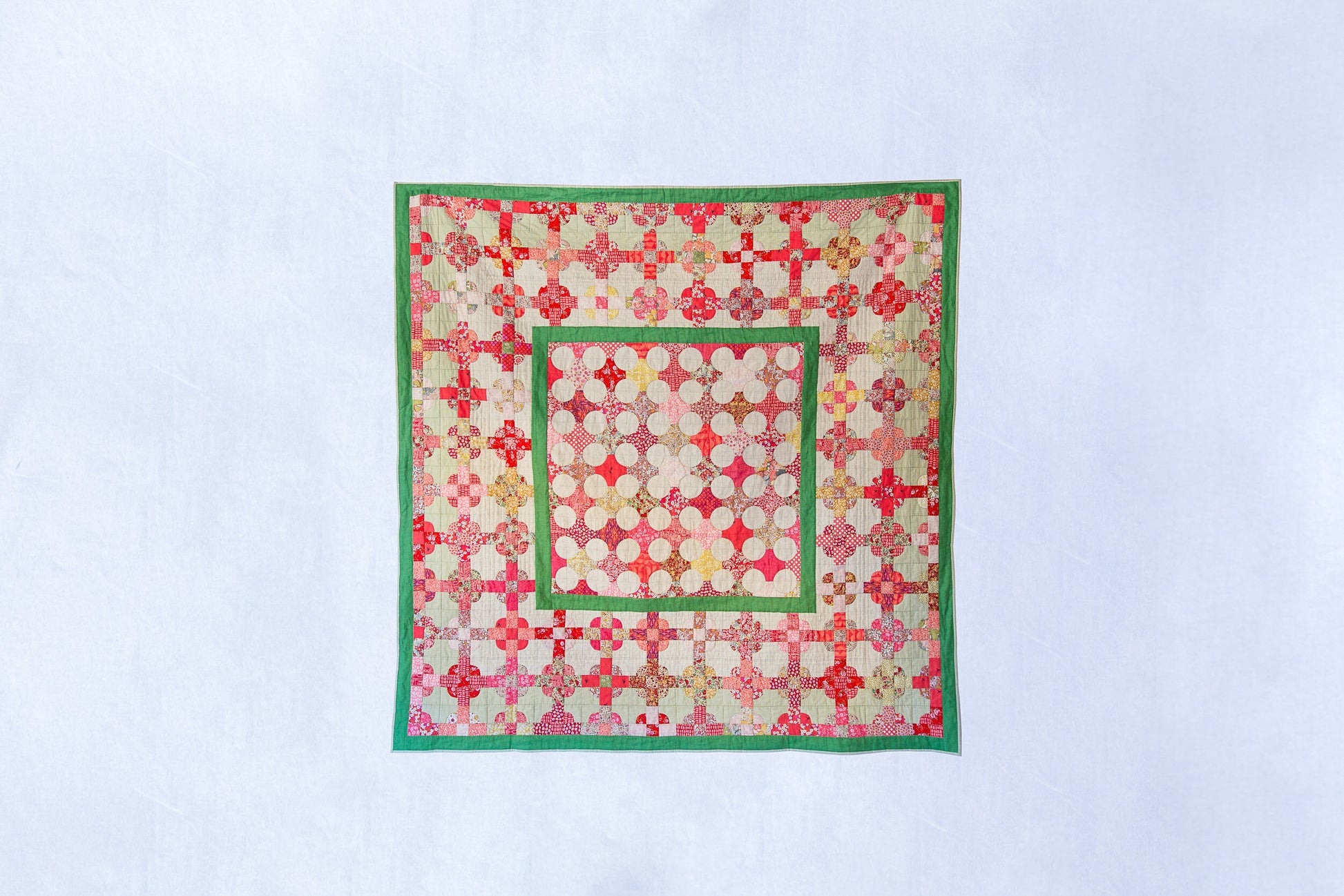 A square quilted textile piece with a geometric patchwork pattern in red, pink, green, and cream, displayed flat against a light blue background.
Photo by MelStoreyPatchwork.com.au [image_link]