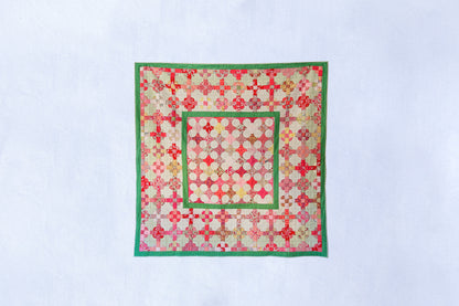 A square quilted textile piece with a geometric patchwork pattern in red, pink, green, and cream, displayed flat against a light blue background.
Photo by MelStoreyPatchwork.com.au [image_link]