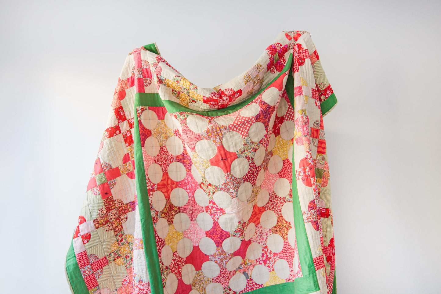 A handmade patchwork quilt draped against a plain white wall. The quilt features a vibrant pattern of red, pink, and orange floral fabrics, large white polka dots on a coral background, and a solid green border.
Photo by MelStoreyPatchwork.com.au [additional_image_link]