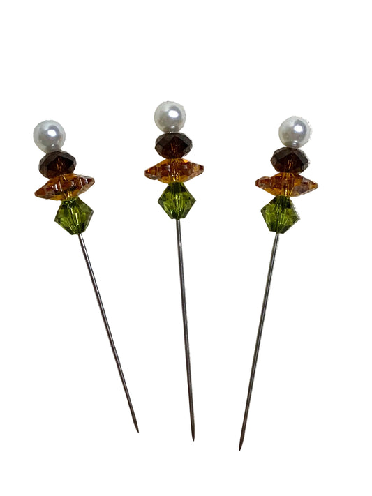 Three decorative hair pins with long dark metal stems, each topped with a white pearl bead, a faceted amber-brown bead, a faceted green bead, and a small faceted dark brown bead, shown on a plain white background.
Photo by TheSurgeonsKnots.com [image_link]