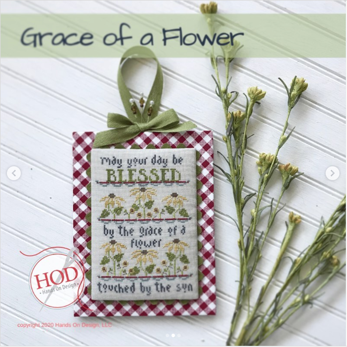 A decorative hanging ornament with a red-and-white gingham border, a green ribbon bow, and embroidered text reading 'May your day be BLESSED by the grace of a flower touched by the sun,' shown on a white paneled background with dried flowers and a 'Grace of a Flower' title banner.
Photo by HandsOnDesign.biz [additional_image_link]