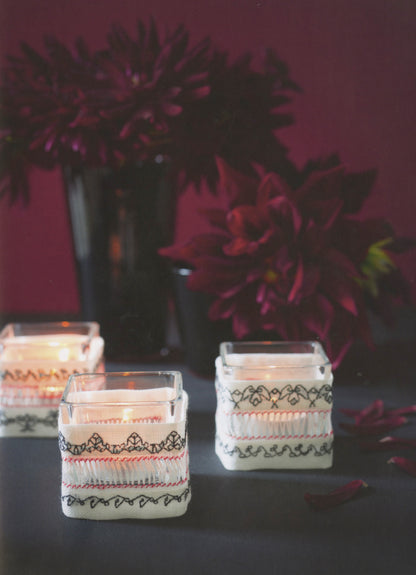 Three square glass candle holders decorated with white fabric wraps featuring black and red embroidered patterns, arranged on a dark table with a dark burgundy floral background.
Photo by Search Press [additional_image_link]
