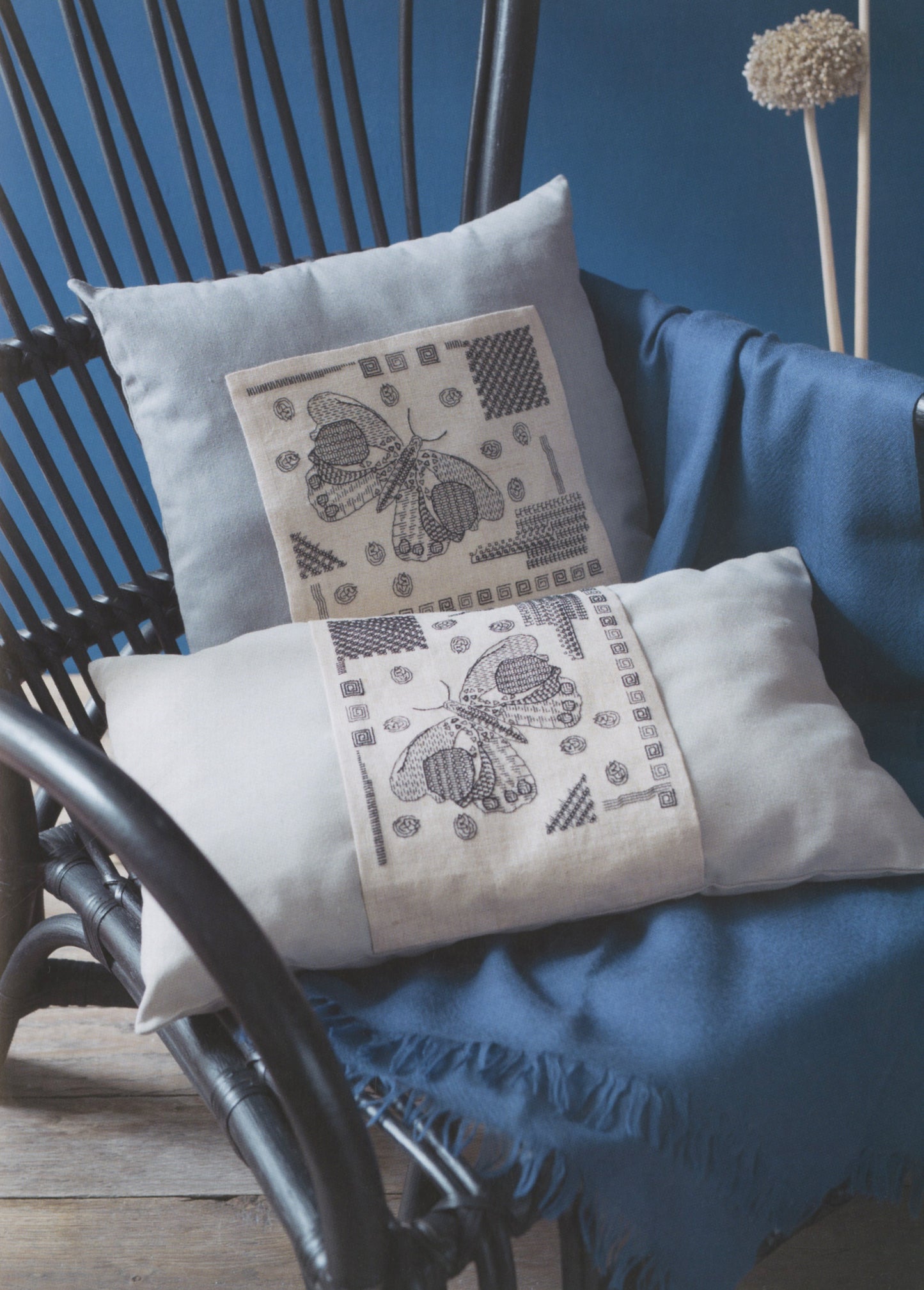Two decorative throw pillows displayed on a blue draped blanket in a dark rattan chair. The pillows are off-white with black geometric and butterfly-like printed patterns.
Photo by Search Press [additional_image_link]