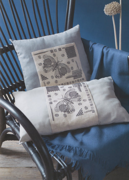 Two decorative throw pillows displayed on a blue draped blanket in a dark rattan chair. The pillows are off-white with black geometric and butterfly-like printed patterns.
Photo by Search Press [additional_image_link]