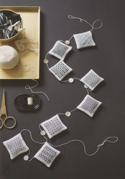 A styled flat-lay of a handmade knitted fabric garland made of small square white pillows with gray and white knit patterns, strung together with white cord and decorated with small round beads. The scene is styled with craft supplies including scissors, a spool of thread, tape, and a gold tray on a dark gray background.
Photo by Search Press [additional_image_link]