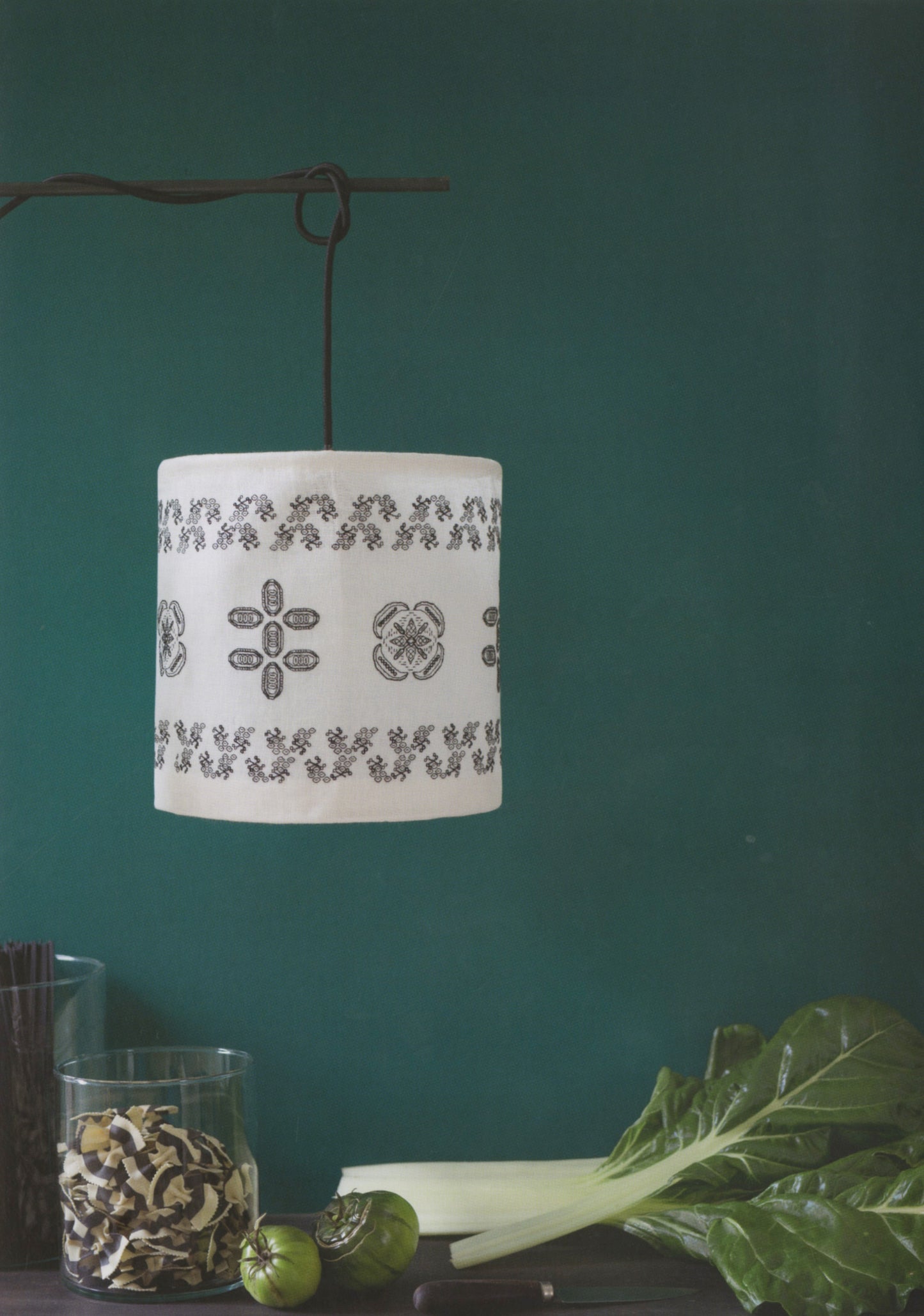 A white fabric drum pendant lamp with black floral and geometric embroidery, hanging from a dark metal rod against a dark teal wall. In the foreground, a glass jar of dried mushrooms, green tomatoes, and leafy greens sit on a dark table.
Photo by Search Press [additional_image_link]
