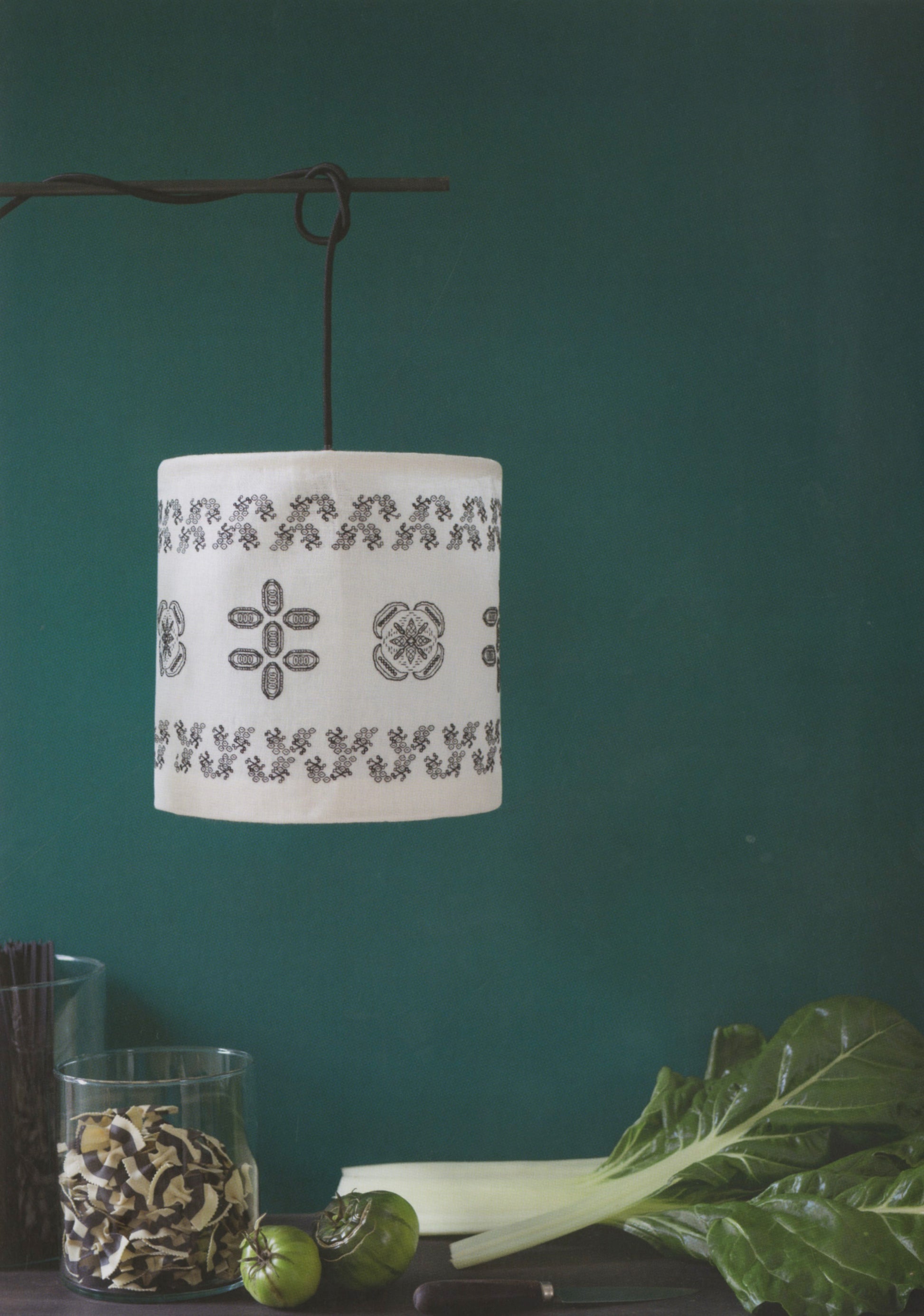 A white fabric drum pendant lamp with black floral and geometric embroidery, hanging from a dark metal rod against a dark teal wall. In the foreground, a glass jar of dried mushrooms, green tomatoes, and leafy greens sit on a dark table.
Photo by Search Press [additional_image_link]