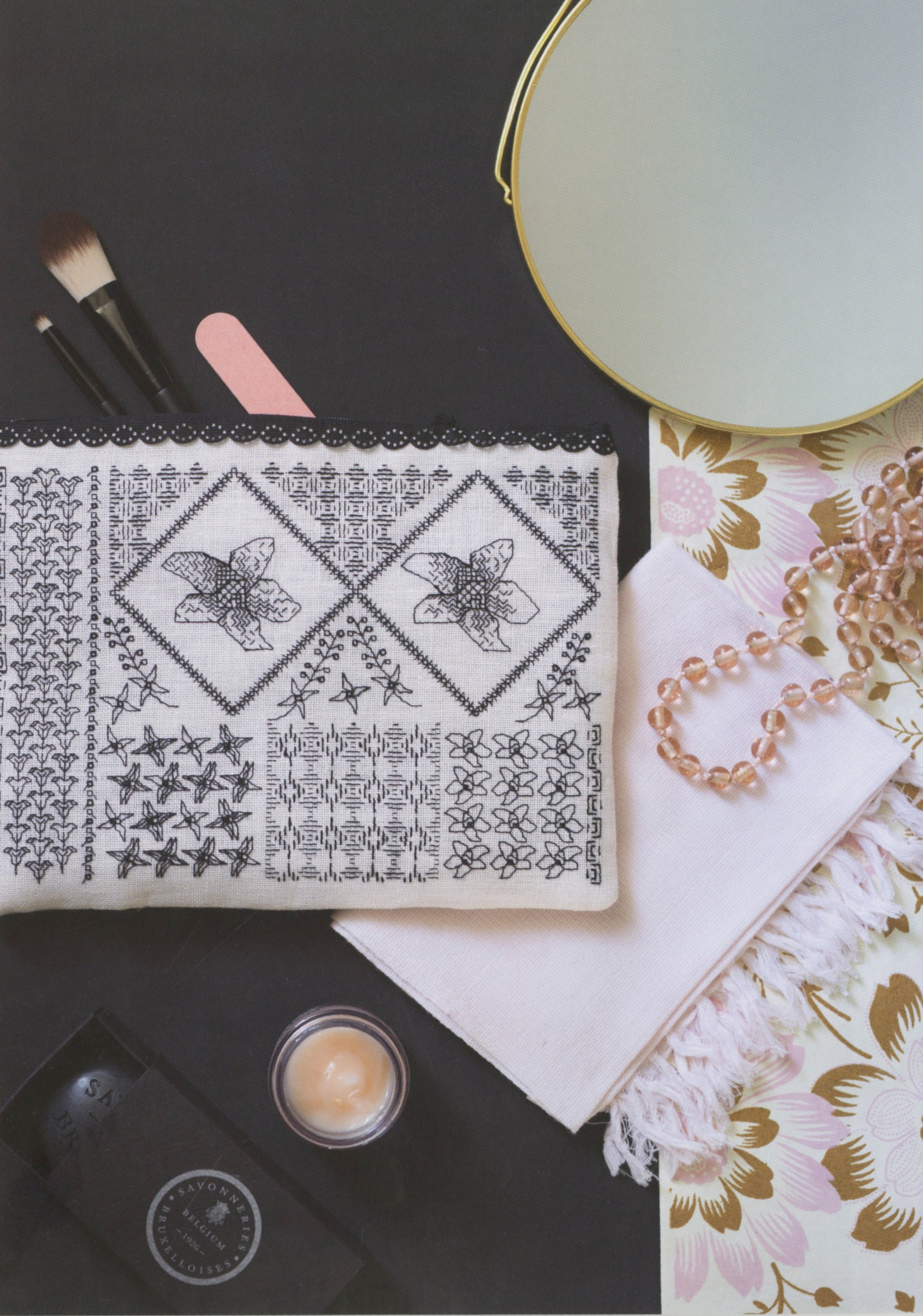 A styled flat-lay featuring a white embroidered cosmetic pouch with black floral and geometric patterns, surrounded by makeup brushes, a pink nail file, a round mirror, a pearl necklace, and a small jar of cream on a dark background.
Photo by Search Press [additional_image_link]