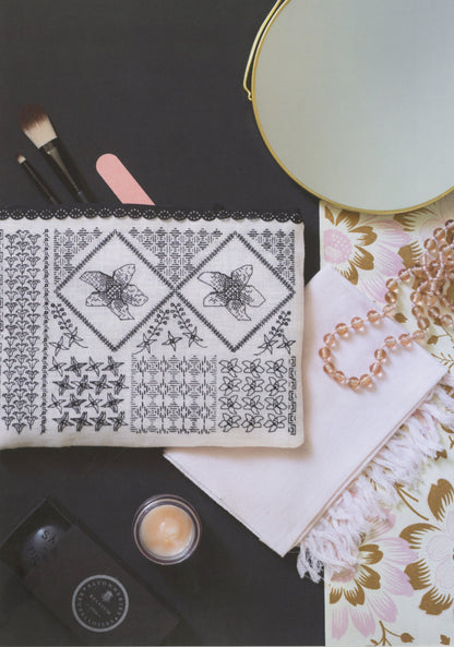 A styled flat-lay featuring a white embroidered cosmetic pouch with black floral and geometric patterns, surrounded by makeup brushes, a pink nail file, a round mirror, a pearl necklace, and a small jar of cream on a dark background.
Photo by Search Press [additional_image_link]