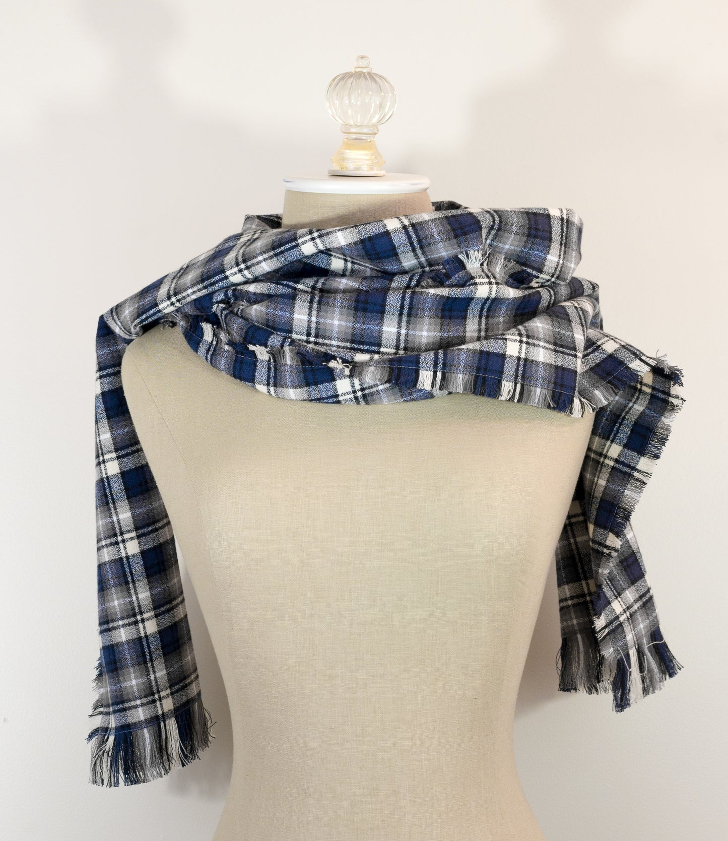 A navy blue, white, and gray plaid scarf with fringe details, draped on a cream-colored mannequin torso against a plain white background.
Photo by Karen King [additional_image_link]
