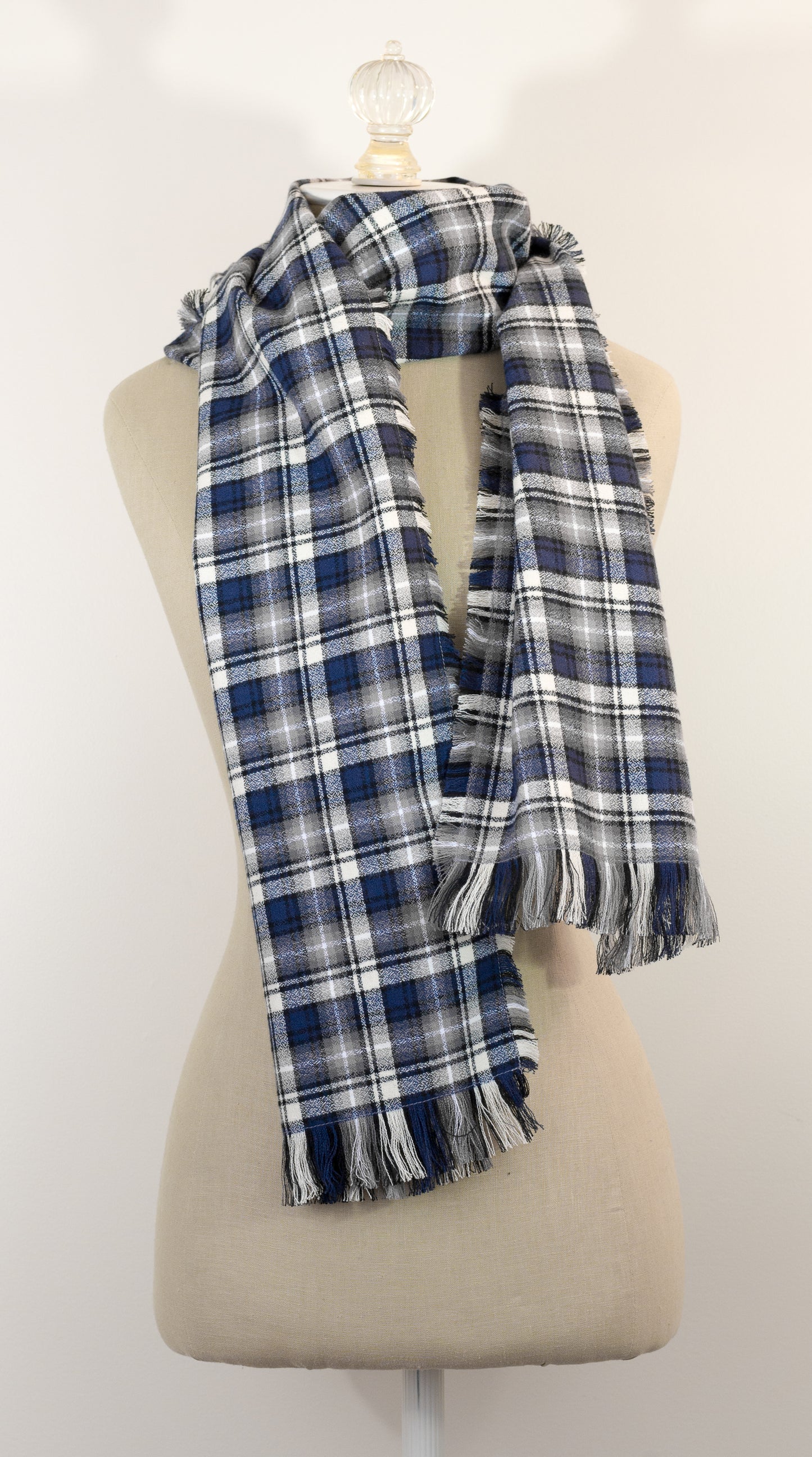 A navy blue, white, and gray plaid scarf with fringe details, draped on a cream-colored mannequin torso against a plain white background.
Photo by Karen King [additional_image_link]