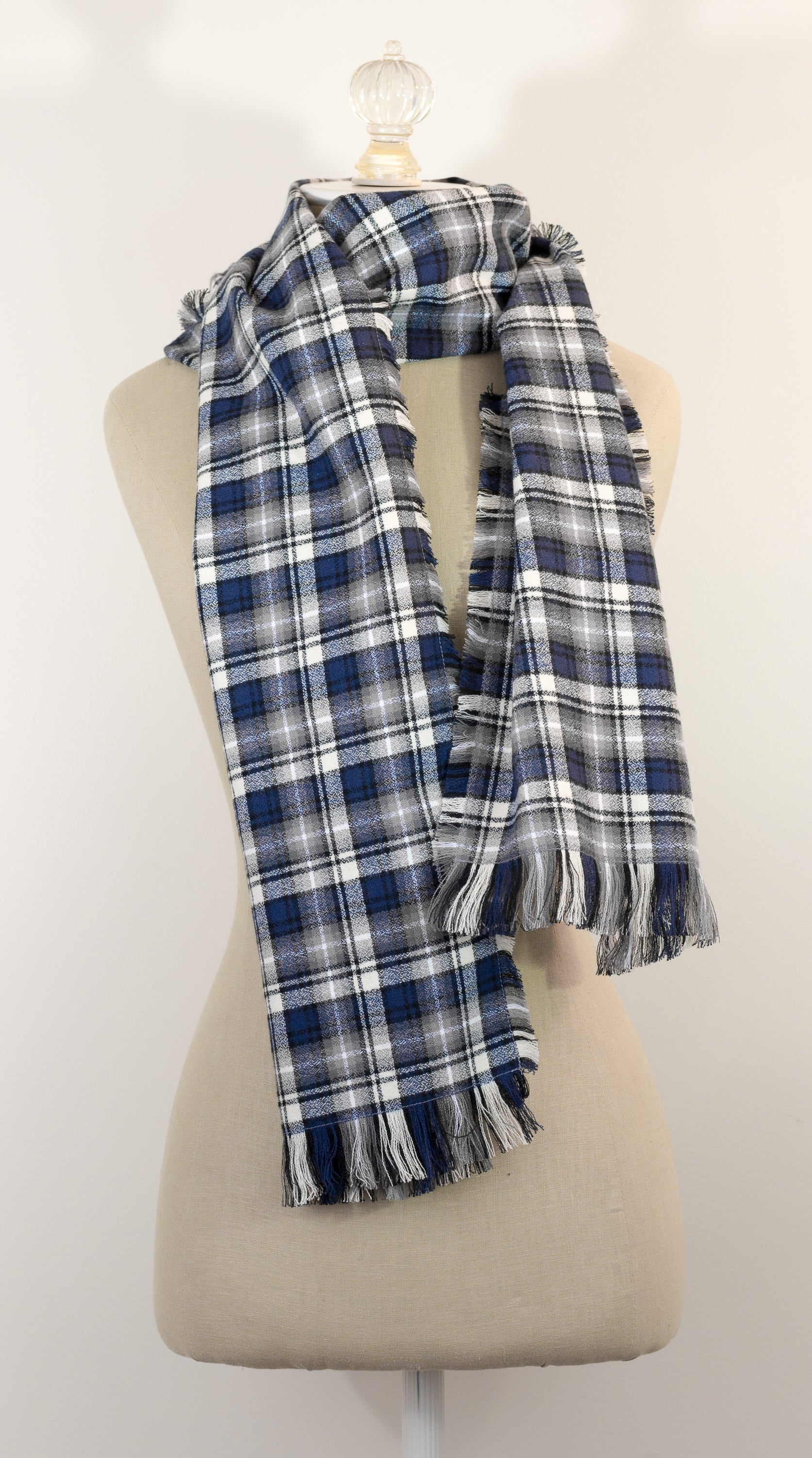 A navy blue, white, and gray plaid scarf with fringe details, draped on a cream-colored mannequin torso against a plain white background.
Photo by Karen King [additional_image_link]