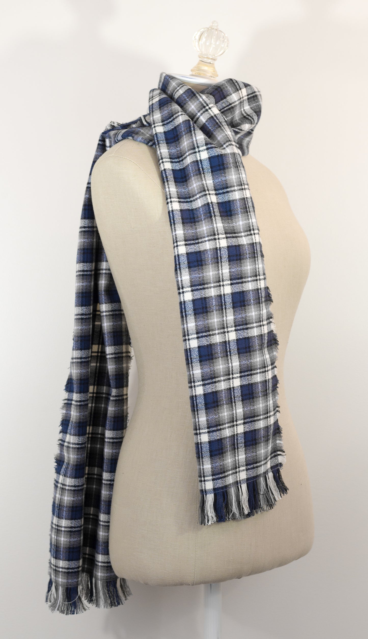 A navy blue, white, and gray plaid scarf with fringe details, draped on a cream-colored mannequin torso against a plain white background.
Photo by Karen King [additional_image_link]