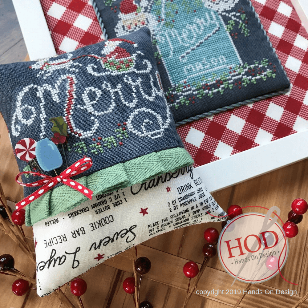 Two decorative cross-stitch pillows displayed on a wooden surface with red-and-white check fabric and holiday garland. The front pillow is cream with black script text, a light green ruffle band, a red polka-dot ribbon bow, and a blue candy embellishment.
Photo by HandsOnDesign.biz [additional_image_link]