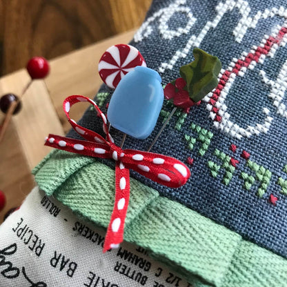 A close-up of a decorative holiday-themed pin cushion with embroidered text, ribbed green fabric, and several pins including a blue oval pin, a red-and-white striped candy pin, and a green holly pin, tied with a red polka-dot ribbon.
Photo by HandsOnDesign.biz [additional_image_link]