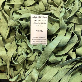 A pile of olive green ribbon or tape strips filling the frame, with a small white paper label placed on top. The label reads 'Body Shot Creations' and 'MERRY'.
Photo by Ladydotcreates.com [additional_image_link]