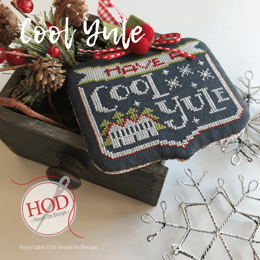 A handmade cross-stitch needlework sample featuring the phrase 'Have a Cool Yule' in white and gray thread on a dark navy fabric background, decorated with red berries, pinecones, and a rustic wooden box. A circular logo for 'Hand On Design' is visible in the lower-left corner.
Photo by HandsOnDesign.biz [image_link]