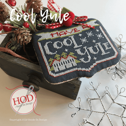 A handmade cross-stitch needlework sample featuring the phrase 'Have a Cool Yule' in white and gray thread on a dark navy fabric background, decorated with red berries, pinecones, and a rustic wooden box. A circular logo for 'Hand On Design' is visible in the lower-left corner.
Photo by HandsOnDesign.biz [image_link]