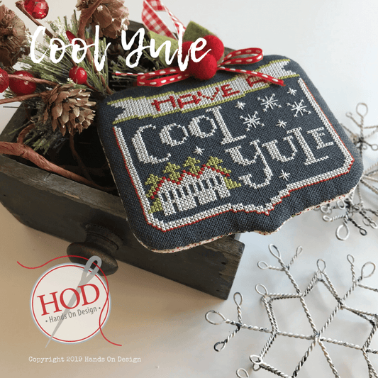 A handmade cross-stitch needlework sample featuring the phrase 'Have a Cool Yule' in white and gray thread on a dark navy fabric background, decorated with red berries, pinecones, and a rustic wooden box. A circular logo for 'Hand On Design' is visible in the lower-left corner.
Photo by HandsOnDesign.biz [image_link]
