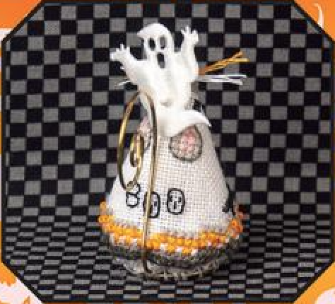 A small Halloween-themed decorative object featuring a white ghost figure above a white cone-shaped candy corn body with orange trim, black embroidered details, and a gold ribbon, set against a black-and-white checkered background.
Photo by JustNan.com [additional_image_link]