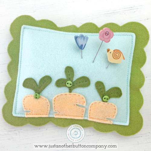 A handmade felt pincushion kit from Just Another Button Company, featuring orange, blue, and green felt pieces, with polymer clay pins topped with various decorative elements, a blue flower, a pink rose and a yellow bird.
Photo by JustAnotherButtonCompany.com [image_link]