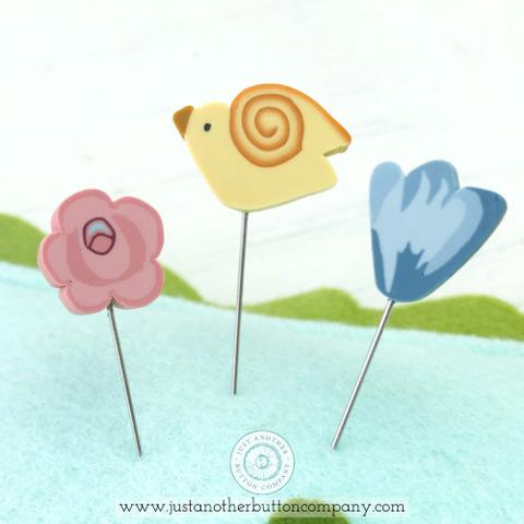 A handmade felt pincushion kit from Just Another Button Company, featuring orange, blue, and green felt pieces, with polymer clay pins topped with various decorative elements. Close up of the pins, one pink rose, one yellow bird and one blue flower.
Photo by JustAnotherButtonCompany.com [additional_image_link]