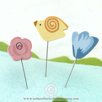 A handmade felt pincushion kit from Just Another Button Company, featuring orange, blue, and green felt pieces, with polymer clay pins topped with various decorative elements. Close up of the pins, one pink rose, one yellow bird and one blue flower.
Photo by JustAnotherButtonCompany.com [additional_image_link]