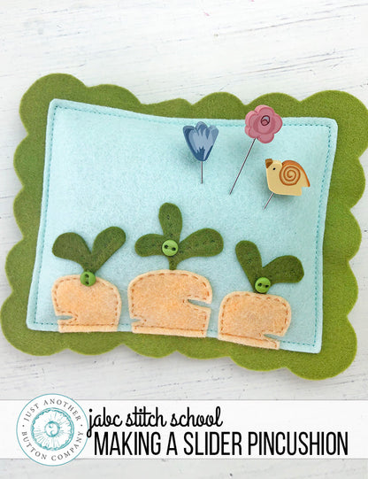 A handmade felt pincushion kit from Just Another Button Company, featuring orange, blue, and green felt pieces, with polymer clay pins topped with various decorative elements, a blue flower, a pink rose and a yellow bird.
Photo by JustAnotherButtonCompany.com [additional_image_link]