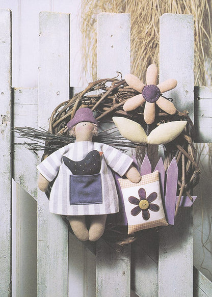 A handmade rustic decorative wreath hanging on a white picket fence. The wreath is made of brown grapevine branches and features a fabric doll wearing a white and purple striped dress, a purple flower, and a small purple garden flag.
Photo by Tildafabrics.com [additional_image_link]