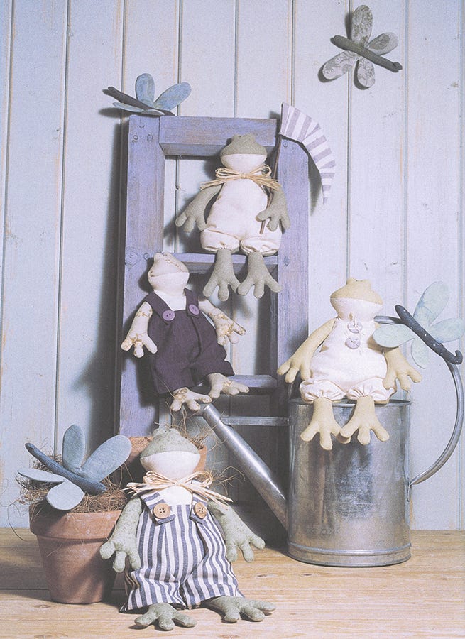 Three fabric frog dolls wearing different outfits are arranged on rustic decorative props, including a wooden ladder, a metal watering can, a potted plant, and a striped fabric tag, set against a light blue paneled wall.
Photo by Tildafabrics.com [additional_image_link]