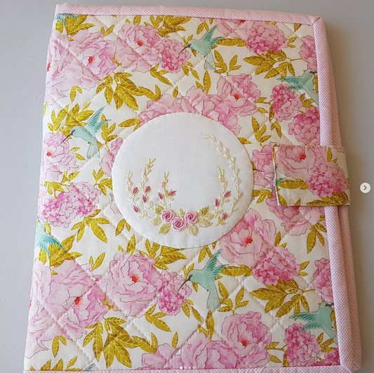 A handmade quilted fabric notebook or journal cover featuring a pink and green floral print with a central round white patch decorated with a floral wreath embroidery. The cover has a pink stitched edge and a matching fabric closure tab on the right side.
Photo by Angela Watson [image_link]