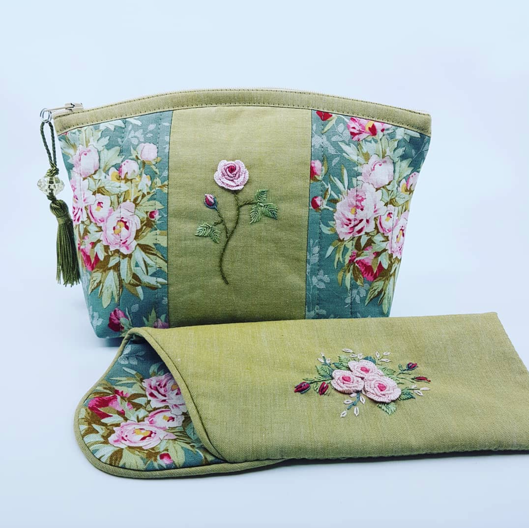 A handmade fabric cosmetic pouch with a matching zippered sleeve, featuring an olive green linen body, floral tapestry panels with pink roses, and embroidered rose motifs on a light blue background.
Photo by Angela Watson [additional_image_link]