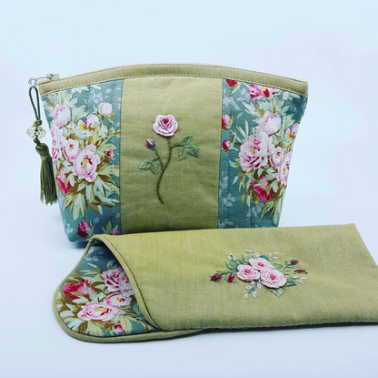A handmade fabric cosmetic pouch with a matching zippered sleeve, featuring an olive green linen body, floral tapestry panels with pink roses, and embroidered rose motifs on a light blue background.
Photo by Angela Watson [additional_image_link]
