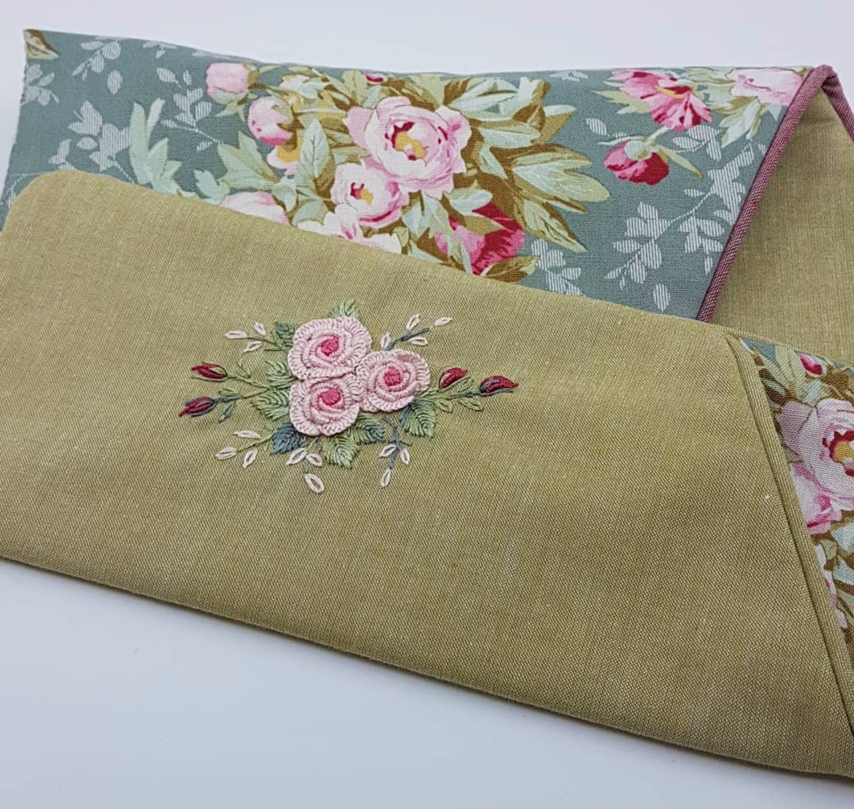 A folded olive green linen fabric piece with embroidered pink roses, shown with another matching floral fabric piece behind it on a white background.
Photo by Angela Watson [additional_image_link]