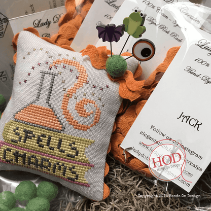 A small embroidered decorative pillow with a spellbook design reading 'Spells Charms' and an orange potion bottle motif, shown with orange ric rac, green felt ball, and printed cards in a styled flat-lay arrangement.
Photo by HandsOnDesign.biz [additional_image_link]