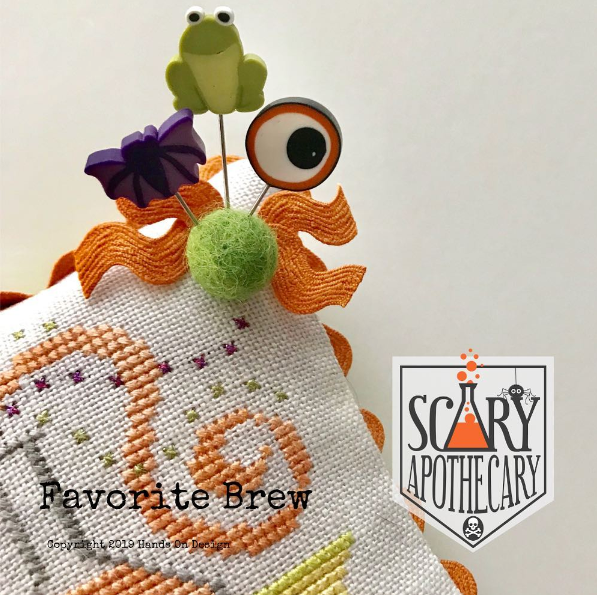 A close-up of a stitched fabric item, likely a cross-stitch sampler or pillow cover, featuring an orange and white abstract design. The fabric is pinned with whimsical Halloween-themed pins including a green frog, a purple bow, a green pom-pom, and an eye on a stick.
Photo by HandsOnDesign.biz [additional_image_link]