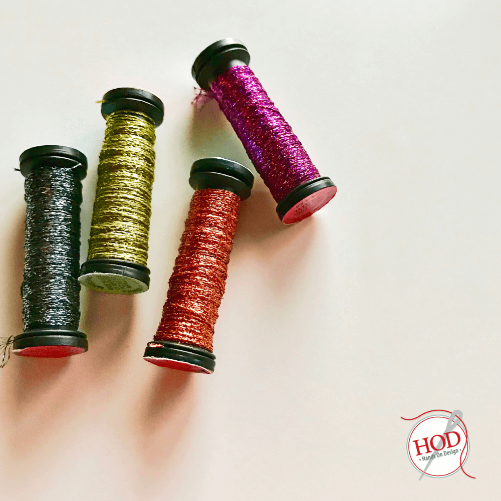 Four decorative spools of glittery thread in black, olive green, red, and magenta, arranged diagonally on a plain off-white background. A circular 'HOD Handmade Designs' logo is visible in the bottom right corner.
Photo by HandsOnDesign.biz [additional_image_link]