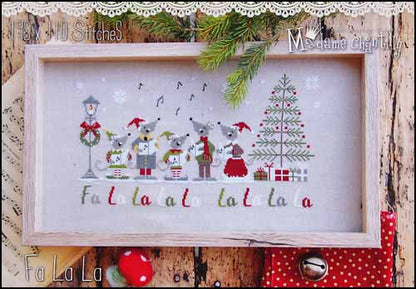 A framed cross-stitch artwork featuring festive woodland animals, a Christmas tree, snowflakes, and the words 'Fa La La' stitched across the bottom. The piece is displayed in a light wood frame against a rustic wood background with lace trim, evergreen branches, and red holiday decorations.
Photo by Madamechantilly.com [image_link]