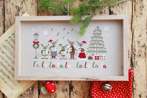 Decorative Christmas scene with cross-stitched design of mice singing around a tree on a wooden background.
Photo by MadameChantilly.com [additional_image_link]