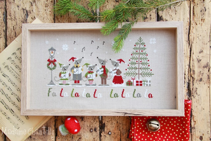 Decorative Christmas scene with cross-stitched design of mice singing around a tree on a wooden background.
Photo by MadameChantilly.com [additional_image_link]