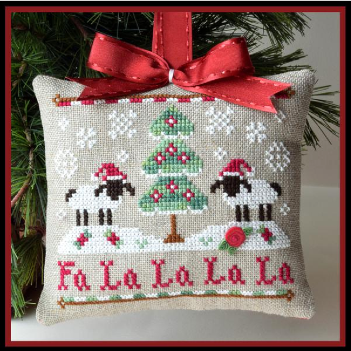 Decorative Christmas pillow with cross-stitch design of sheep, trees, and 'Fa La La La La' text, hanging on a tree.
Photo by Country Cottage Needleworks [image_link]