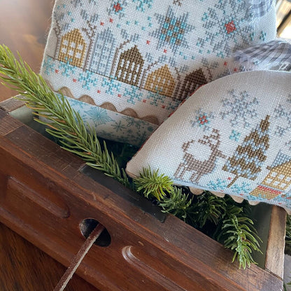 Two decorative cross-stitch pillows with winter holiday designs, including snowflakes, houses, and a deer and tree motif, styled in a wooden box with green pine branches.
Photo by HandsOnDesign.biz [additional_image_link]