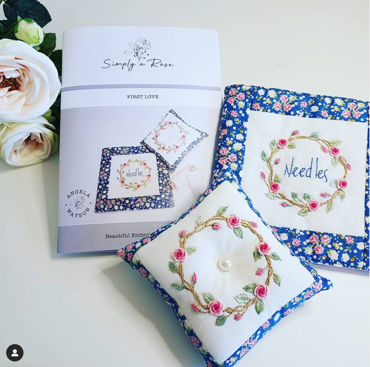 A styled flat-lay of a sewing pattern booklet titled 'Simply Rose First Love' next to two finished embroidered pillow sham designs. The pillows feature white centers with pink rose wreaths and blue floral borders, set against a clean white background with decorative white roses on the left.
Photo by Angela Watson [image_link]