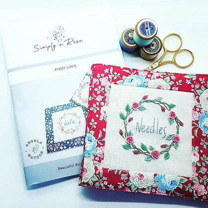 A handmade fabric journal cover with a floral red border and a central cream patch embroidered with a green wreath and the word 'Needles', shown with a printed booklet, spools of thread, and gold scissors on a white background.
Photo by Angela Watson [additional_image_link]